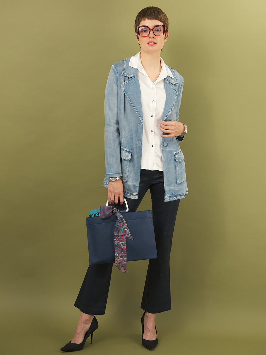 Woman holding a blue bag with a patterned tie wearing a white aliferous shirt and jane jacket by inlarkin against a green background