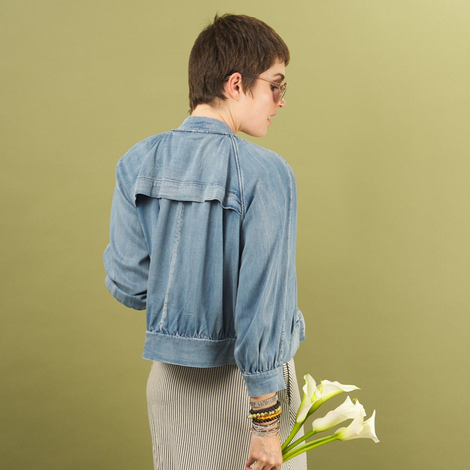 Woman holding white flowers wearing a denim bomber jacket by inlarkin showing the back detail against a green background