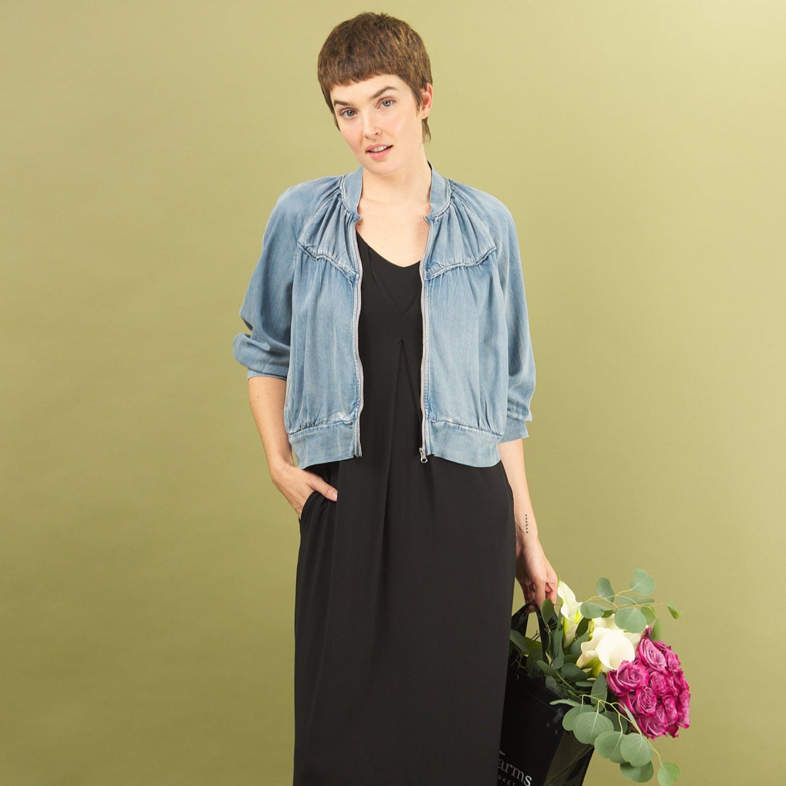 Woman in a black dress and denim boy bomber jacket by inlarkin holding flowers against a green background