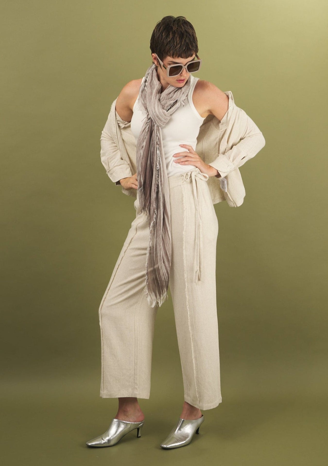 Woman wearing a linen fray pant and shirt by inlarkin with a beige scarf, and silver shoes on a green background