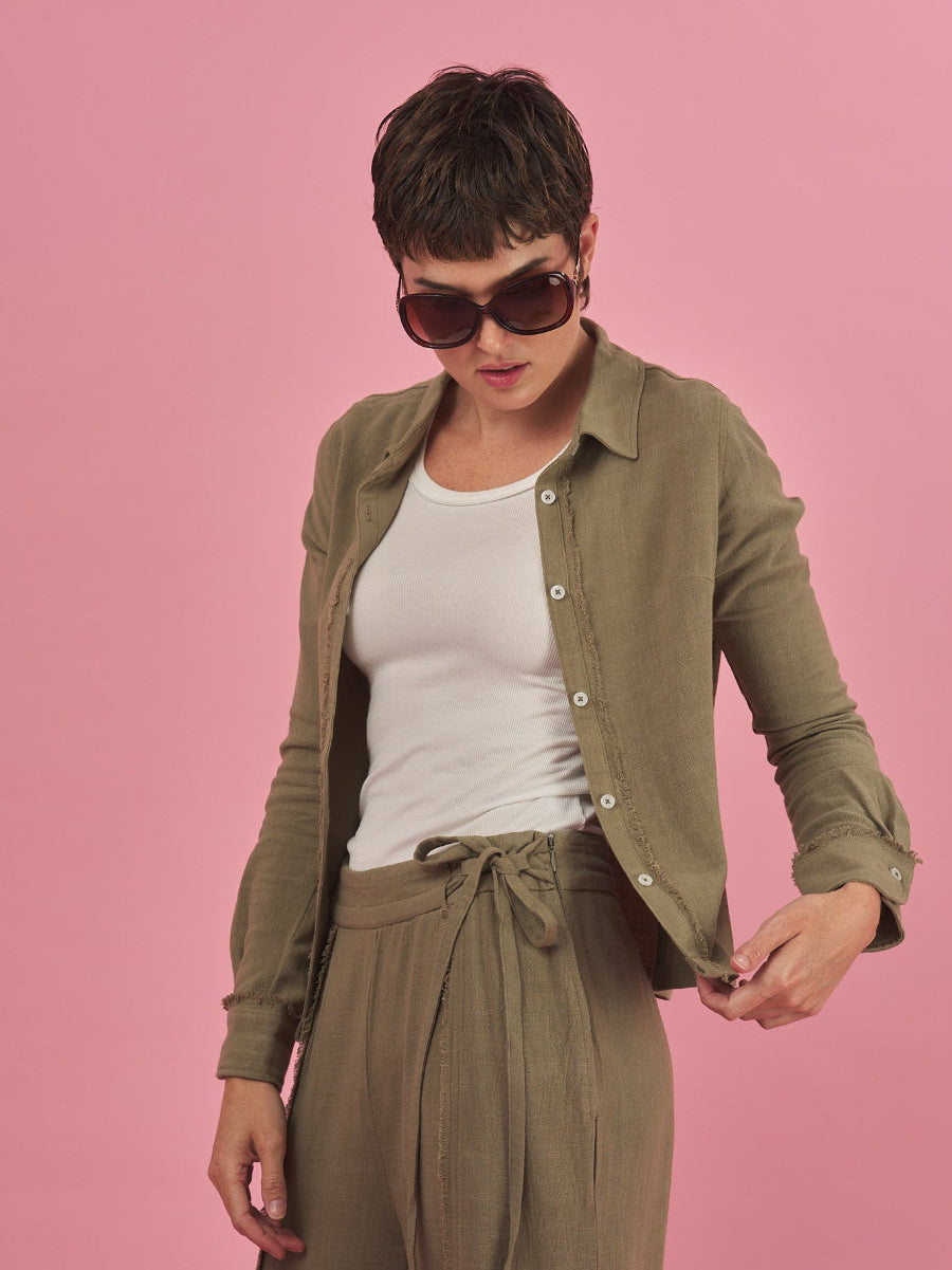 Woman wearing a sage green linen shirt, white tank top and sage green linen pants by inlarkin against a pink background