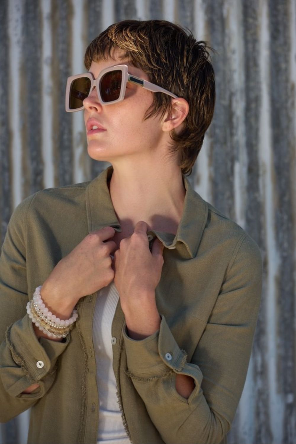 woman wearing sunglasses and a sage green linen shirt against a corrugated metal background