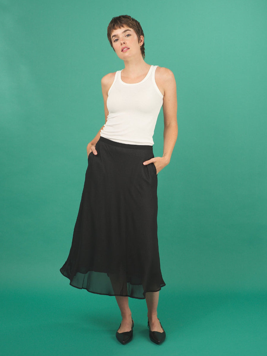 Woman wearing a white tank top and black slay skirt by inlarkin against a green background