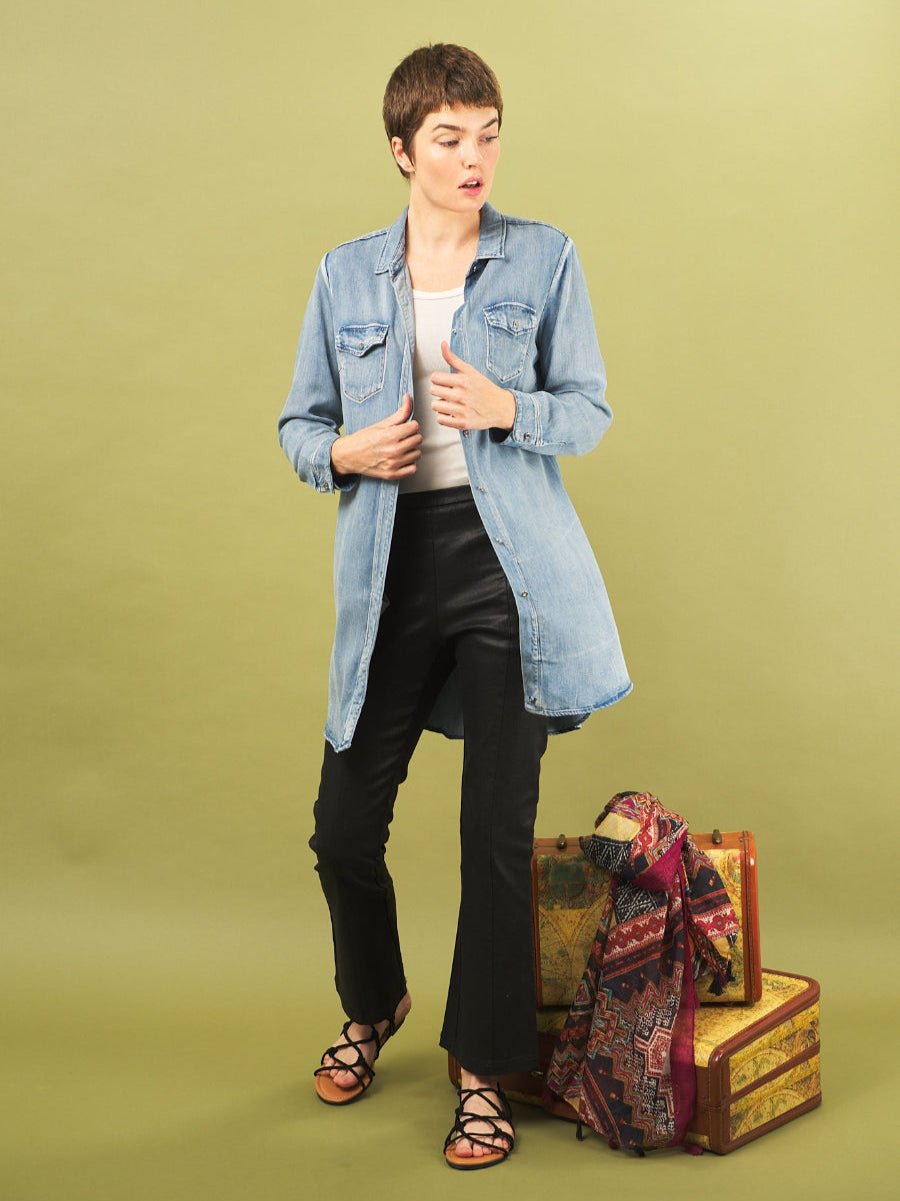 Woman in a sy denim shirt by inlarkin and black pants standing next to vintage suitcases on a green background