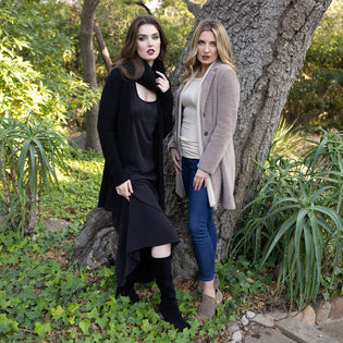  Two women standing in front of a tree in a lush garden setting wearing outfits by designer mary beth larkin, including the unreal jean 2.0 and wingspan dress with cashmere cardigans from the inlarkin clothing line