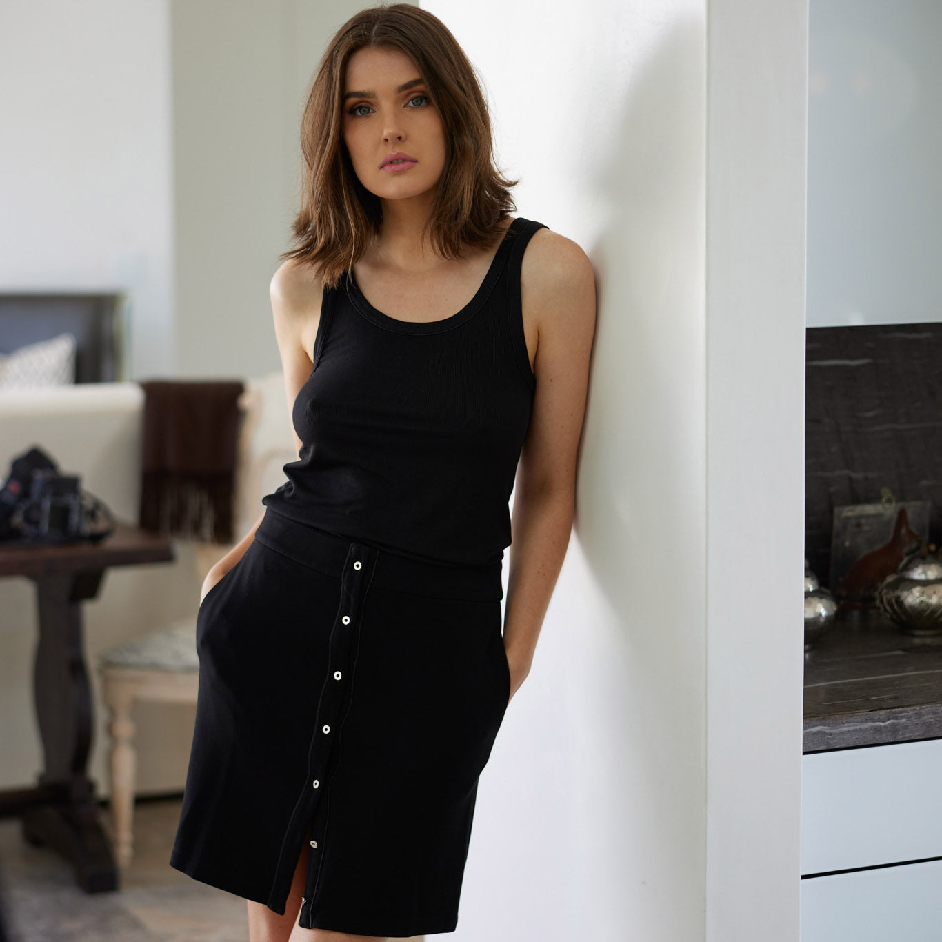 woman standing against a white wall in a contemporary beach house wearing a black variegated tank top and black button up skirt by inlarkin