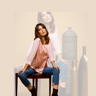  model is sitting on chair wearing a pink button up blouse and jeans 