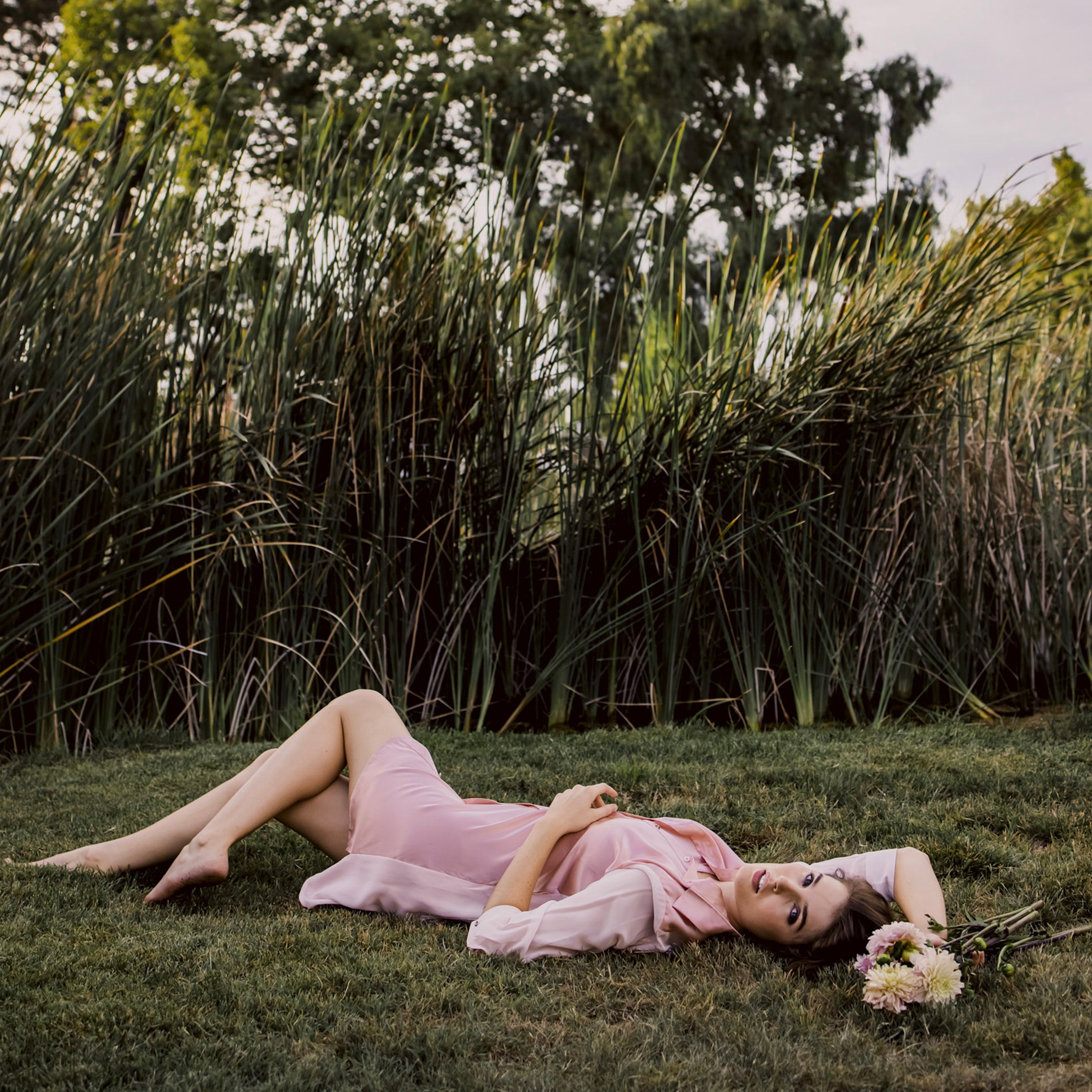 Model wearing pink aliferous shirt dress by inlarkin lying on green grass holding a bouquet of flowers