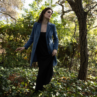  woman in lush greenery outdoors wearing a black long wingspan dress with a longer royal blue blazer over the top of it showing a transitional outfit for winter