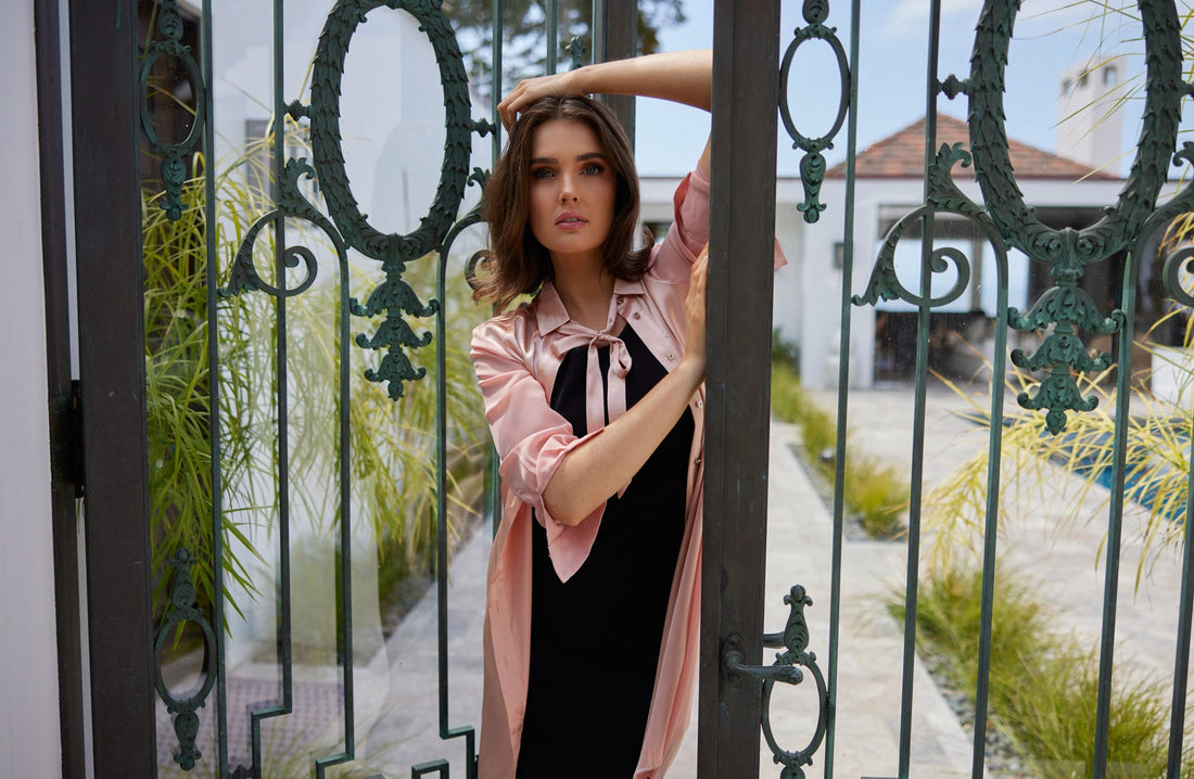  woman standing at decorative metal gates wearing a black form fitting mariposa dress and coral colored long aliferous shirt dress over with a matching ribbon tied at the neck, all clothing by inlarkin