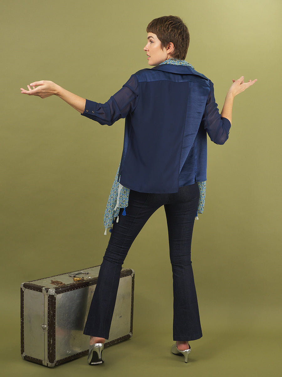 Woman in a navy aliferous shirt by inlarkin standing with arms outstretched on a green background with a piece of luggage