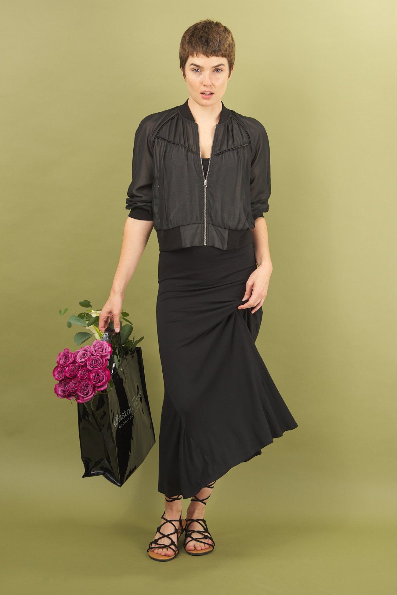 Woman in black sharyn dress and beauty bomber jacket by inlarkin holding a black bag with pink flowers on a green background