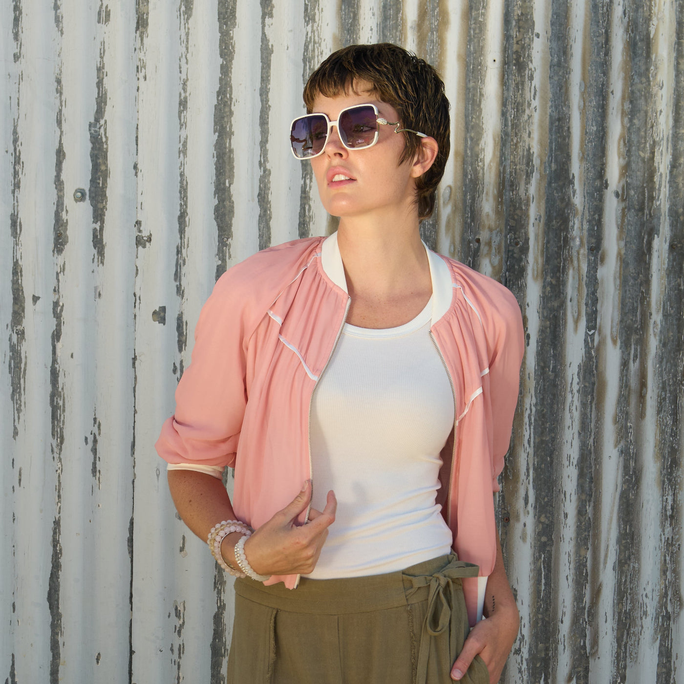 Woman wearing a coral beauty bomber jacket by inlarkin, white top, and green sage linenpants against a corrugated metal wall.