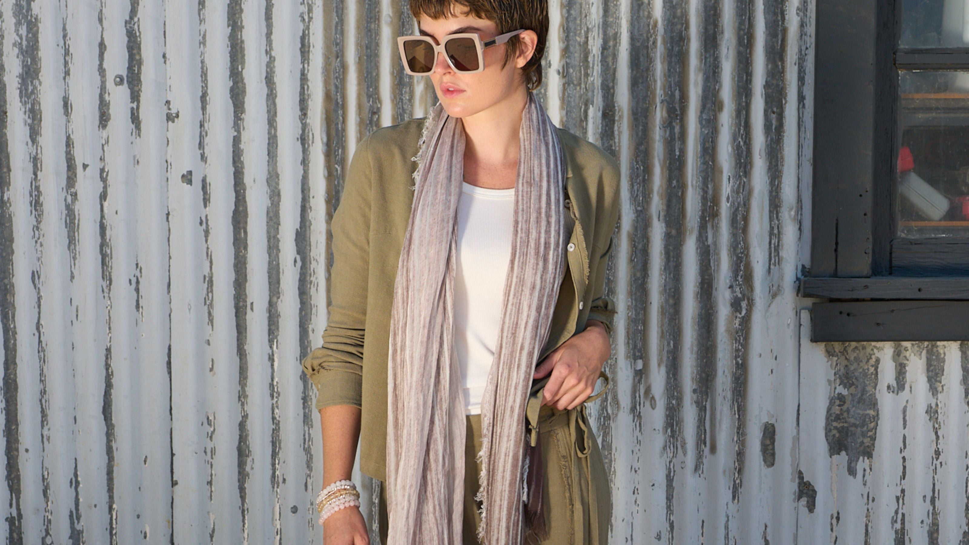 Woman wearing sunglasses, linen fray pant and shirt by inlarkin standing against a corrugated metal wall