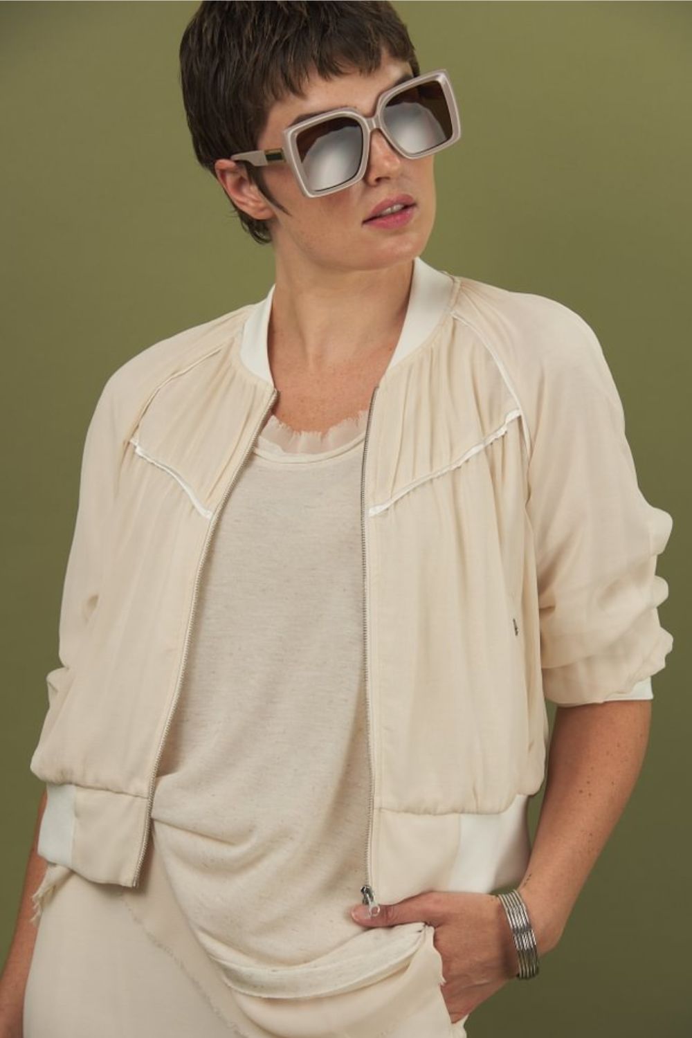 woman wearing the glasswing shell 2.0 in cream paired with a beauty bomber jacket in cream by inlarkin against a green backdrop