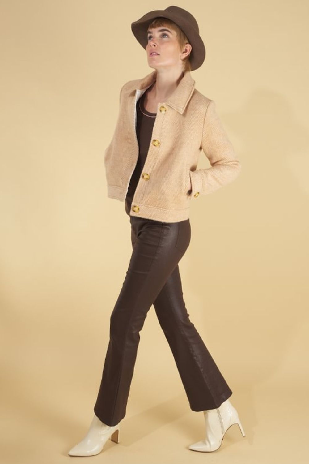 Woman wearing a herringbone harriet jacket by inlarkin, brown hat, and dark pants on a beige background