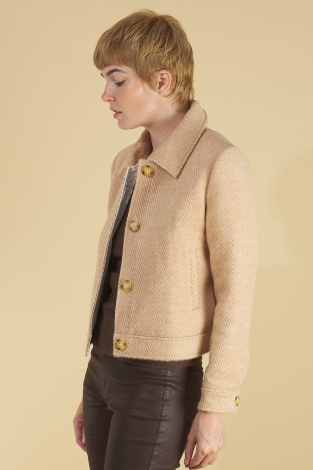 Woman wearing a herringbone harriet jacket by inlarkin, brown hat, and dark pants on a beige background