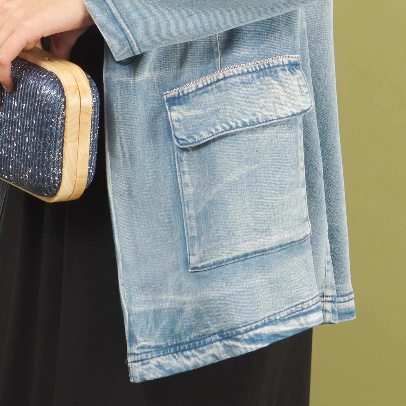 Woman wearing a jane denim jacket by inlarkin with beading along the pocket holding a blue glittery clutch against a green background