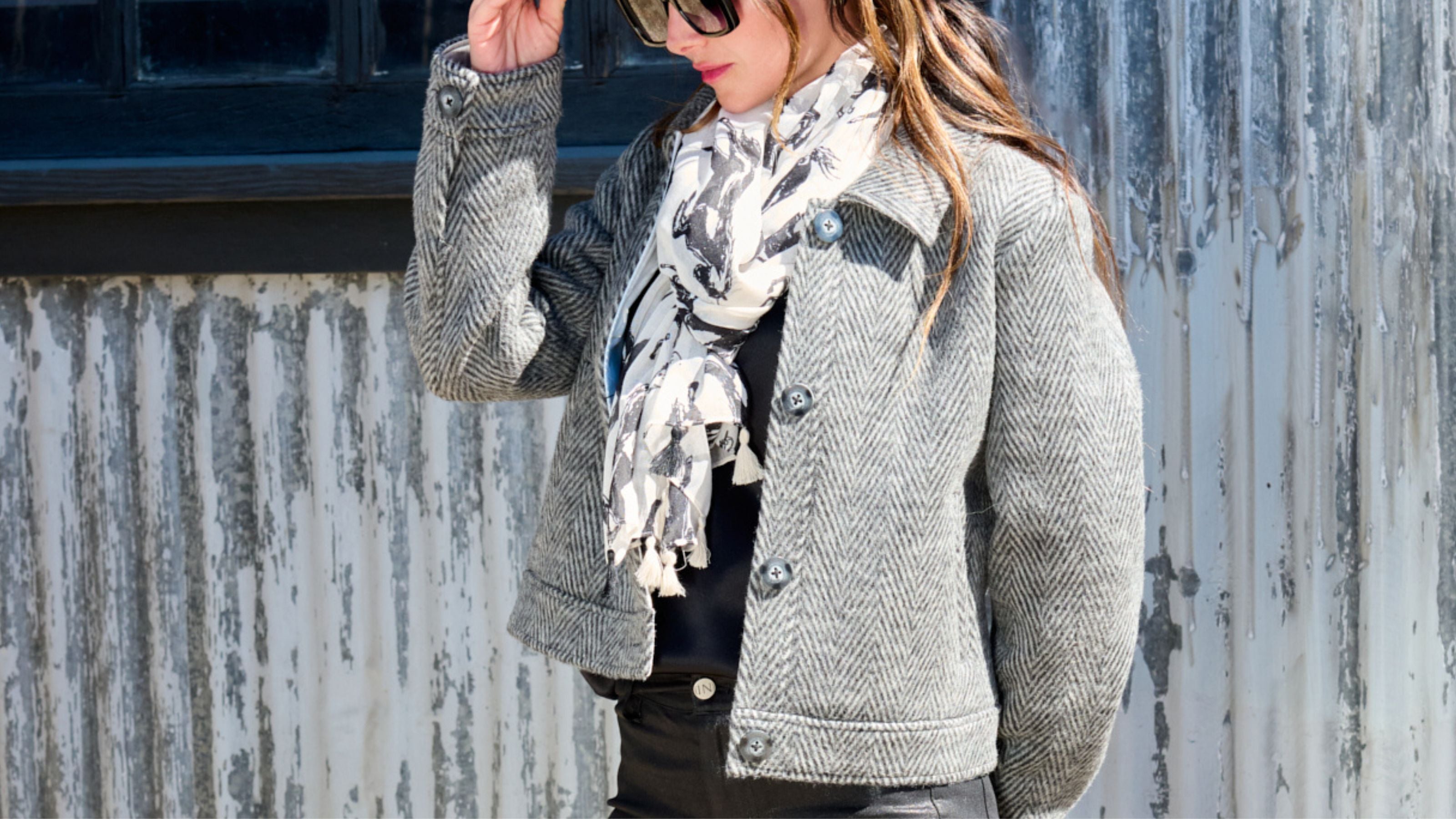 Woman wearing a black herringbone harriet jacket, black curve gloss jeans by inlarkin and a patterned scarf with a black cowboy hat against a textured wall.