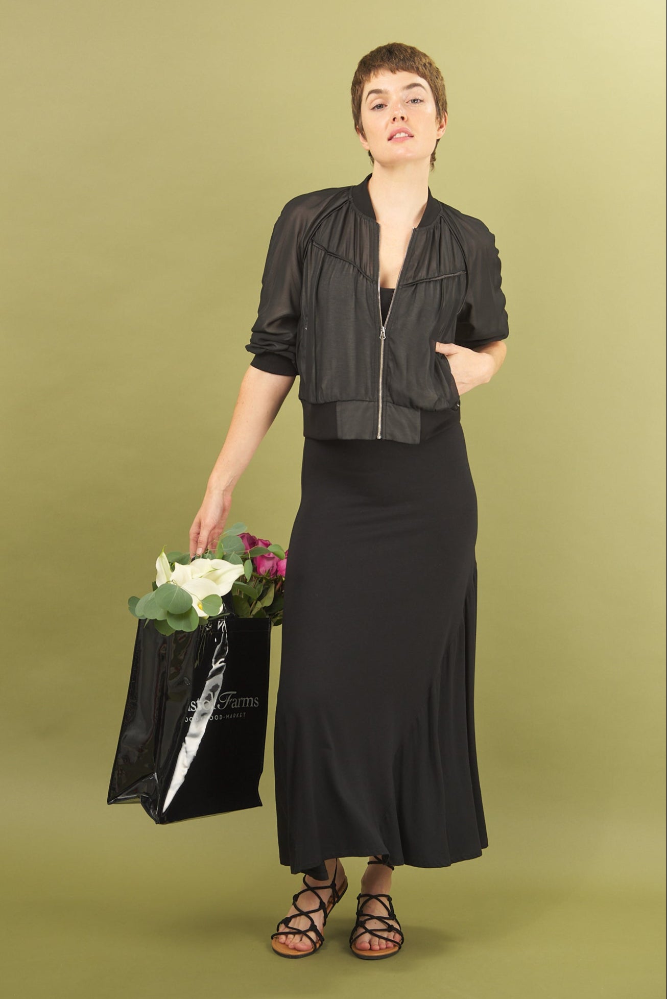 Woman in black inlarkin sharyn dress and beauty bomber jacket outfit holding a black bag with flowers against a green background