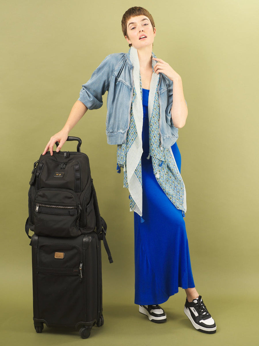 Woman in a cobalt blue sharyn dress by inlarkin with a denim boy bomber jacket holding a black suitcase on a green background