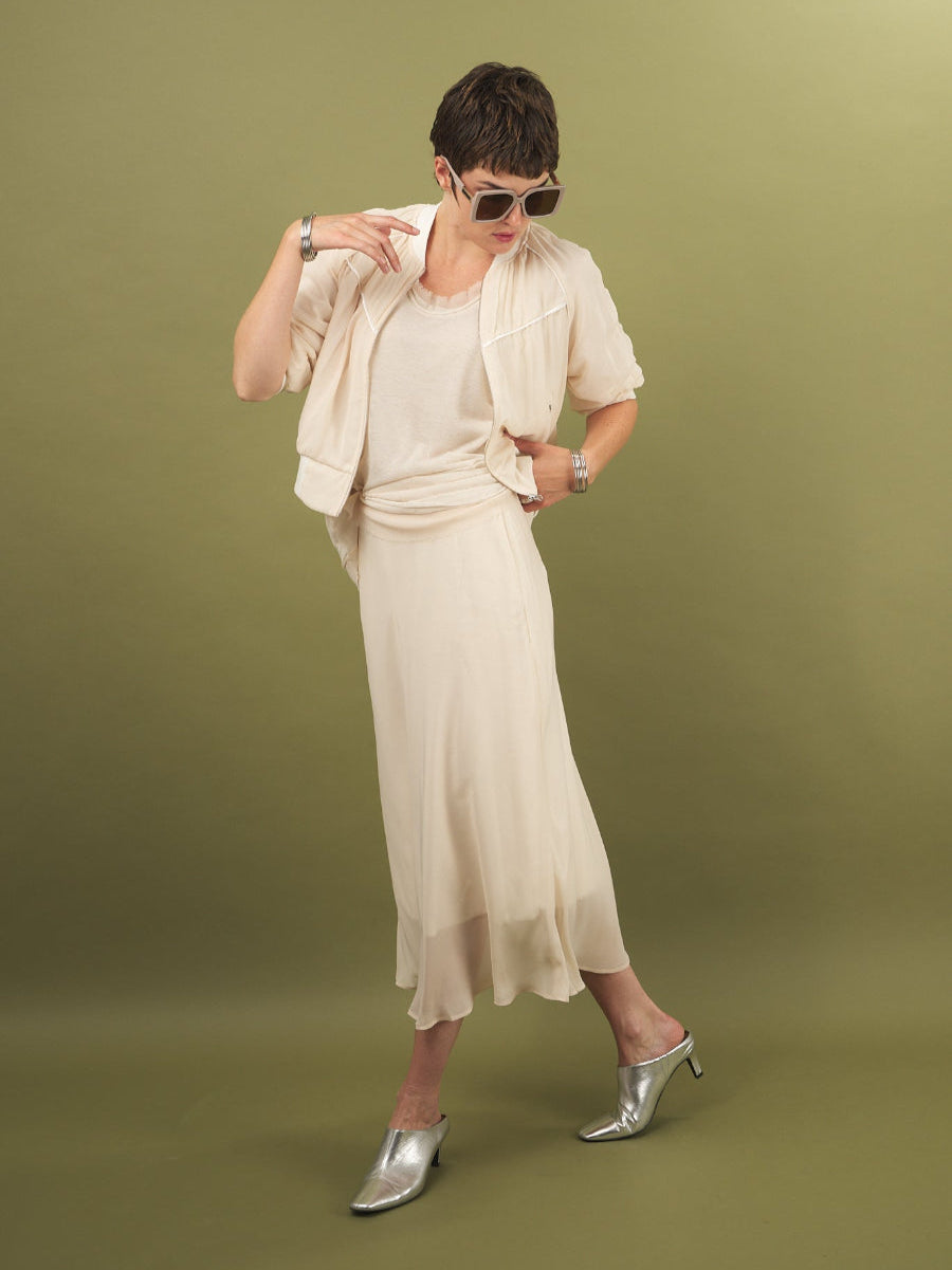Woman in a cream slay skirt and beauty bomber jacket outfit by inlarkin with sunglasses on a green background