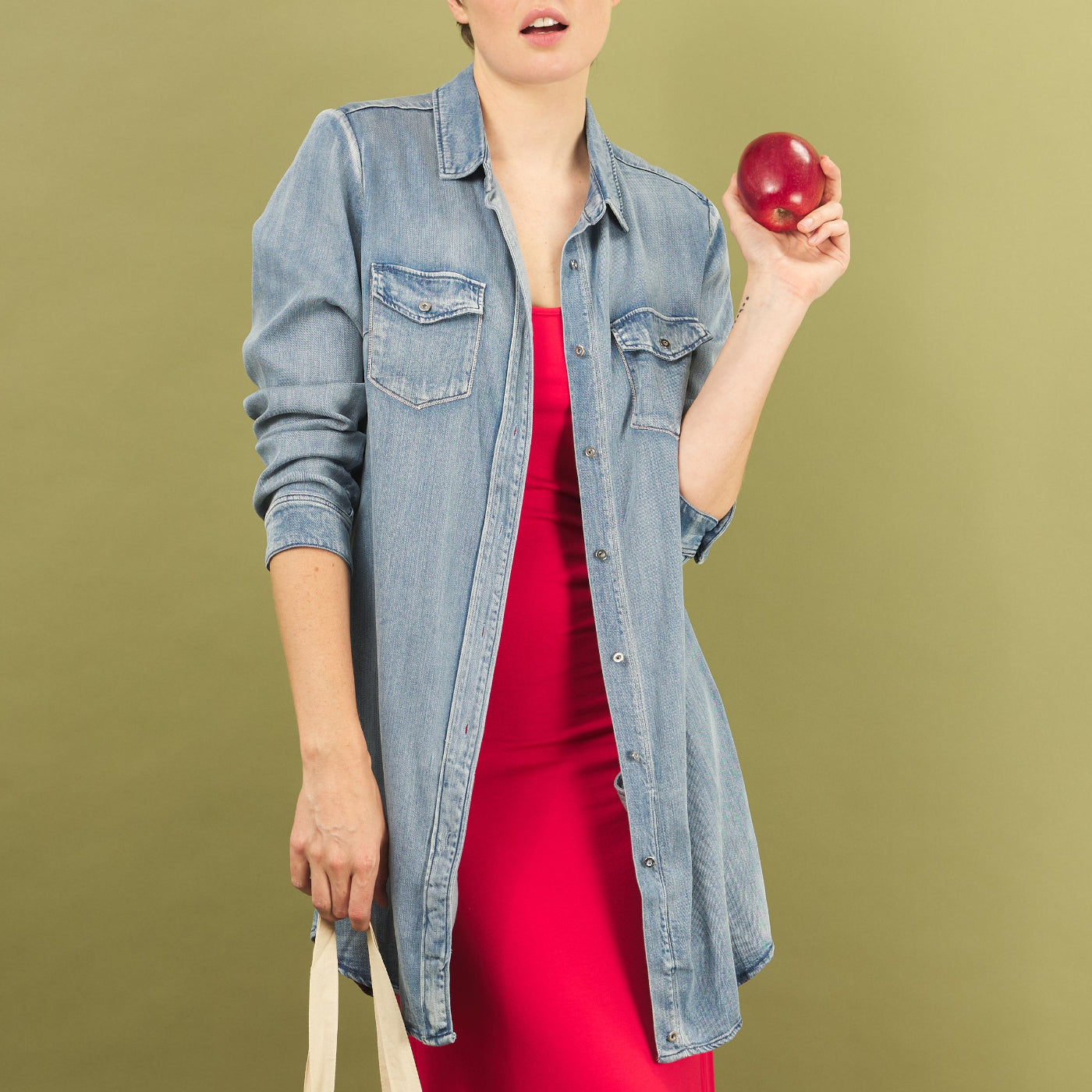 Woman wearing a denim sy shirt by inlarkin over a red sharyn dress, holding a red apple against a green background