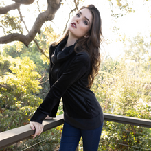  woman standing on a deck among the trees wearing a black engineered cowl sweatshirt by inlarkin showing the detailed cowl neckline and pockets