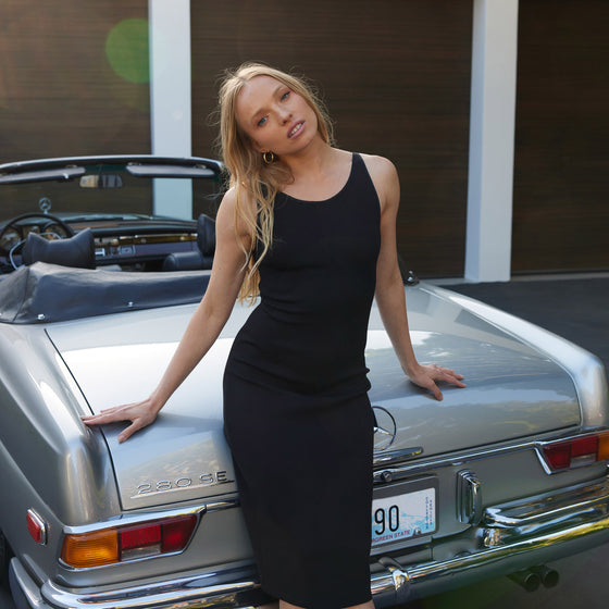model leaning against the back of a vintage gray mercedes benz convertible wearing a form fitting black mariposa dress by inlarkin